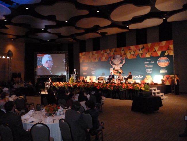 Premiação do Gestor Eficiente da Merenda Escolar, na imagens há mesas com cadeiras à esquerda para o centro com alguns convidados nelas e ao fundo, um palco decorado com flores à frente, com alguns participantes nele. No fundo do palco há um grande painel com o desenho do menino maluquinho com a panela na cabeça, grafo e faca na mão, pronto para saborear a comida que está na mesa com um copo de leite. Do lado do painel há uma tela onde é exibida a filmagem do evento, para ser visto por pessoas que estavam longe do palco.